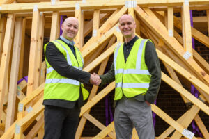 Two men wearing hi-vis shaking hands in front of a some timber roof trusses all stacked together. The two men are celebrating a business acquisition - Taylor Lane Timber Frame today announces new ownership following an acquisition by Cala Group, a UK home builder
